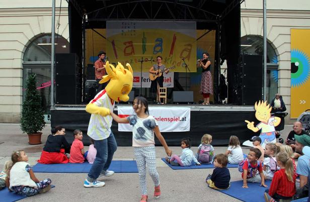 Kinder sitzen vor einer Bühne, während eine Band spielt und ein Maskottchen tanzt.