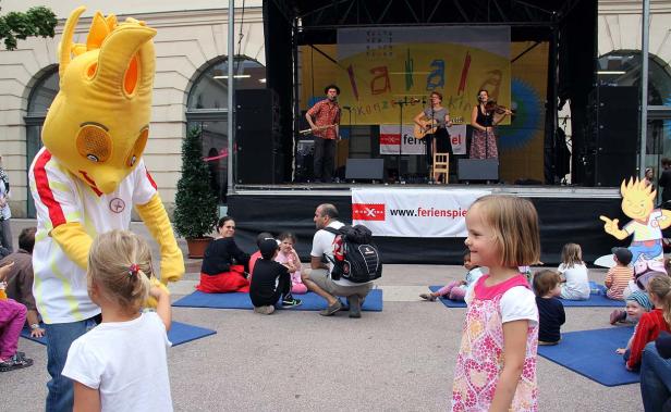 Kinder sitzen vor einer Bühne, auf der eine Band spielt; ein gelbes Maskottchen steht daneben.