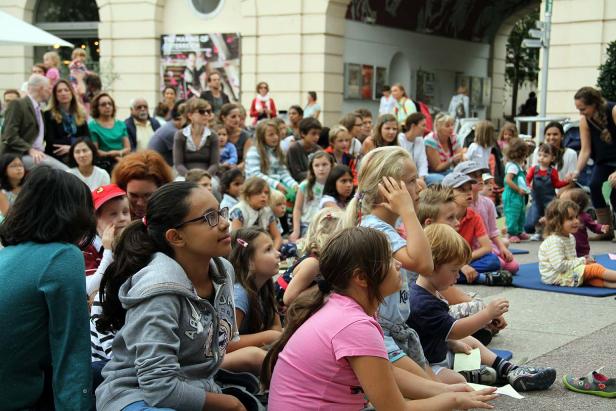 Eine Gruppe von Kindern und Erwachsenen sitzt im Freien und schaut aufmerksam zu.