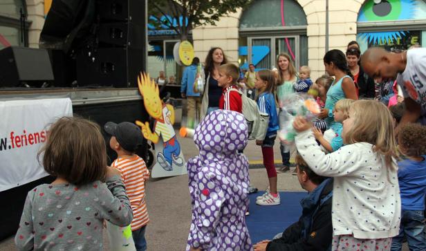 Eine Gruppe von Kindern und Erwachsenen versammelt sich vor einer Bühne.