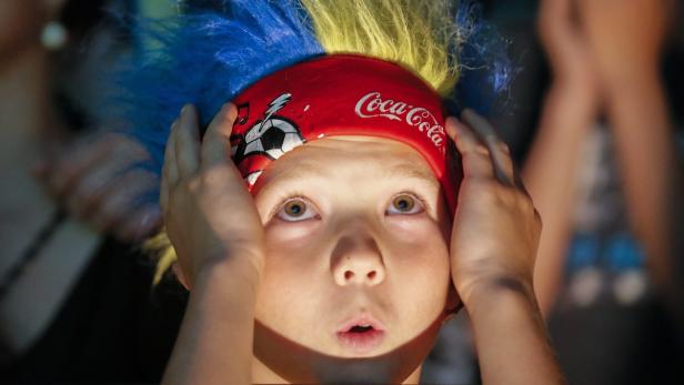 Ein Kind mit blau-gelber Perücke und Coca-Cola-Stirnband schaut überrascht nach oben.