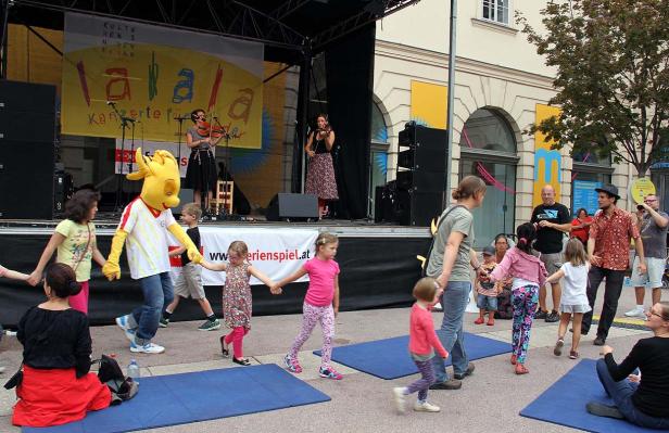 Kinder tanzen mit einem Maskottchen vor einer Bühne mit Musikern.