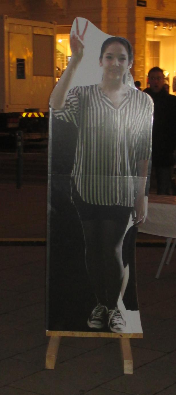 Ein Pappaufsteller einer jungen Frau mit erhobenem Victory-Zeichen steht auf einem Gehweg.