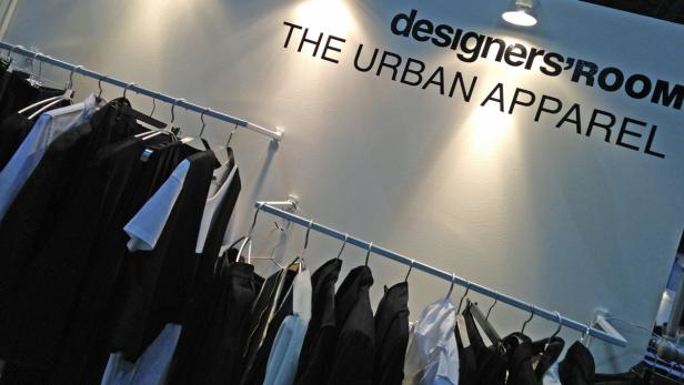 Kleidungsstücke hängen an einer Stange vor einer Wand mit der Aufschrift „designers'ROOM THE URBAN APPAREL“.