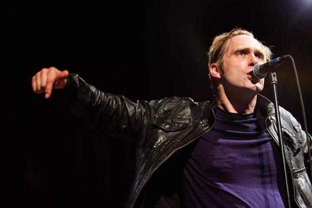 Ein Mann mit Lederjacke singt in ein Mikrofon und zeigt mit dem Finger.