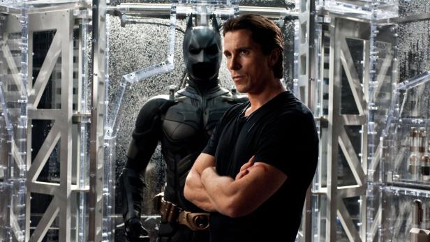 Christian Bale steht neben seinem Batman-Anzug in einer Vitrine.