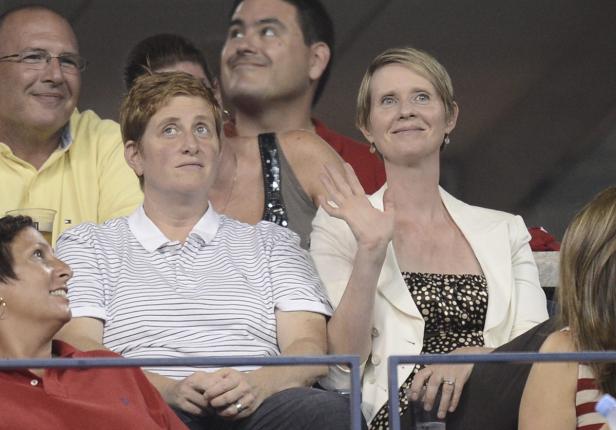 Cynthia Nixon sitzt mit anderen Zuschauern auf einer Tribüne und winkt.