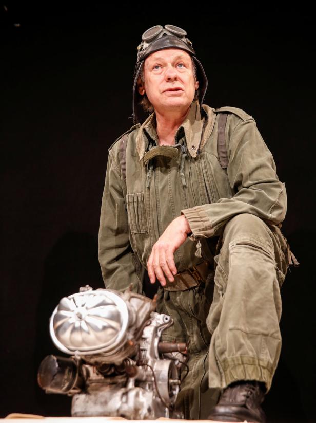 Ein Mann in Fliegerkluft kniet neben einem Flugzeugmotor.