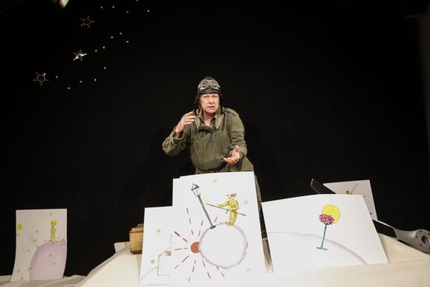 Ein Mann in Fliegerausrüstung präsentiert Zeichnungen aus „Der kleine Prinz“.