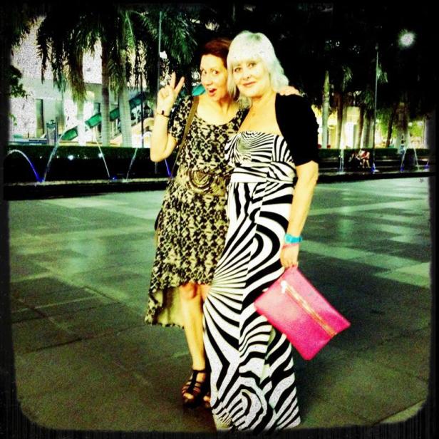 Zwei Frauen posieren vor Palmen und einem Springbrunnen für ein Foto.