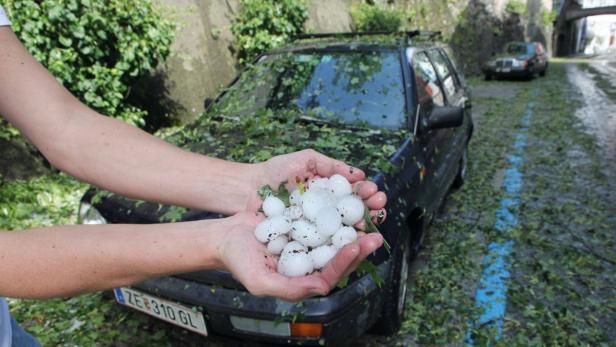 Eine Person hält eine Handvoll Hagelkörner vor einem von Hagel bedeckten Auto.