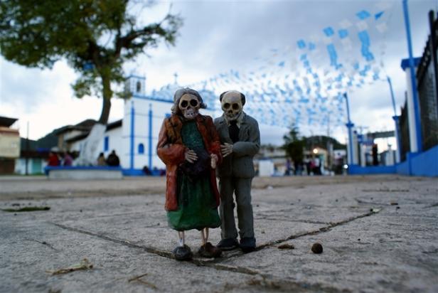 Zwei Skelettfiguren stehen auf einem Platz vor einer Kirche, geschmückt mit blauen Girlanden.