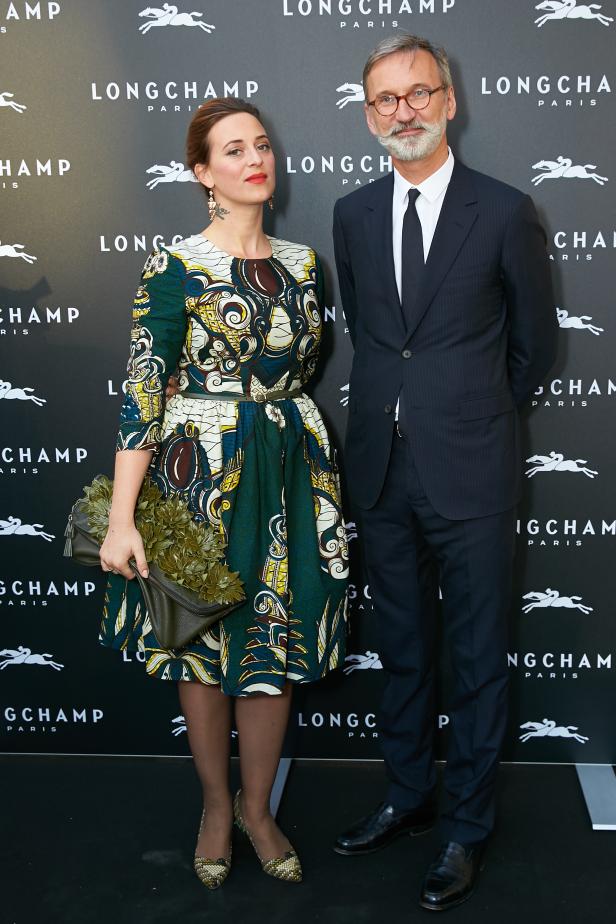 Eine Frau in einem gemusterten Kleid und ein Mann im Anzug posieren vor einer Longchamp-Wand.