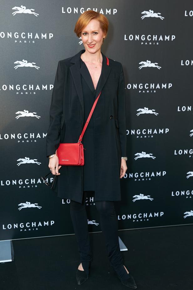 Eine Frau mit rotem Haar posiert vor einer Longchamp-Wand mit einer roten Handtasche.
