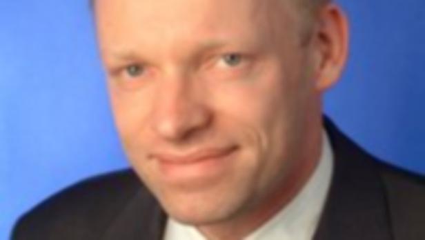 Ein Mann mit hellem Haar und einem dunklen Anzug vor einem blauen Hintergrund.