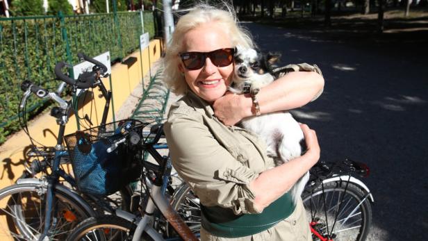 Eine Frau mit Sonnenbrille umarmt einen kleinen Hund neben Fahrrädern.