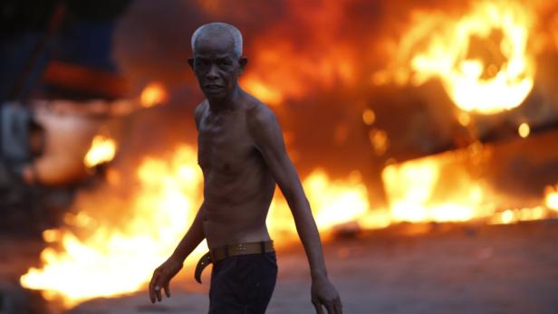 Ein älterer, barfüßiger Mann steht vor einem großen Feuer.