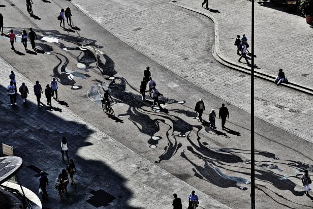 Eine belebte Straße mit Fußgängern und einem Radfahrer, deren Schatten auf ein Kunstwerk fallen.
