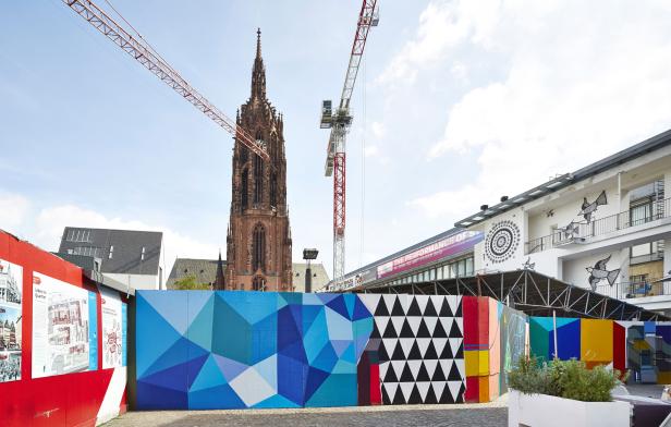 Die Frankfurter Paulskirche hinter einer bunten Bauzaun-Installation mit zwei Kränen im Bild.