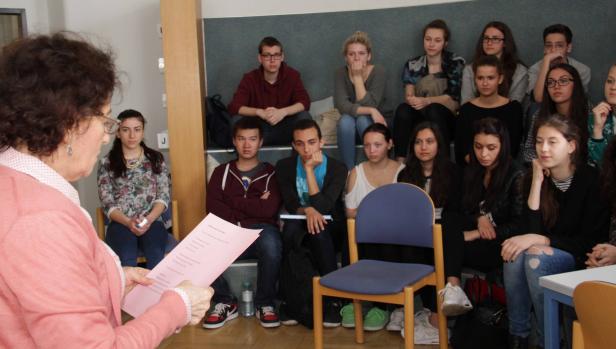 Eine Lehrerin liest vor einer Gruppe von Schülern aus einem rosafarbenen Blatt Papier vor.