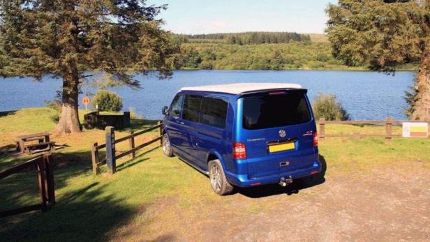 Ein blauer VW-Bus steht an einem Seeufer mit Bäumen im Hintergrund.