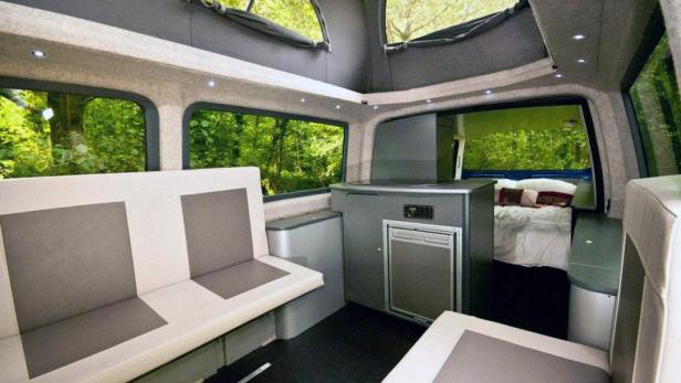 Innenansicht eines Wohnmobils mit Sitzecke, Küchenzeile und Bett im Hintergrund.