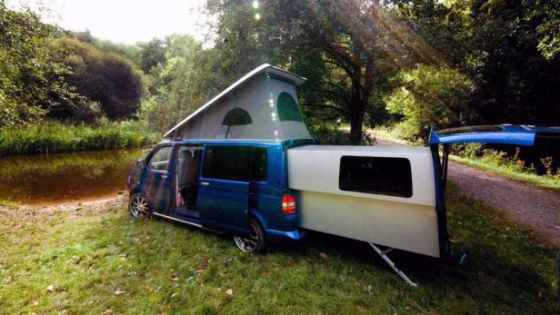 Ein blauer VW-Camper mit Aufstelldach steht auf einer Wiese am Ufer eines Flusses.