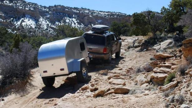 Ein Geländewagen zieht einen silbernen Anhänger auf einem steinigen Weg in den Bergen.