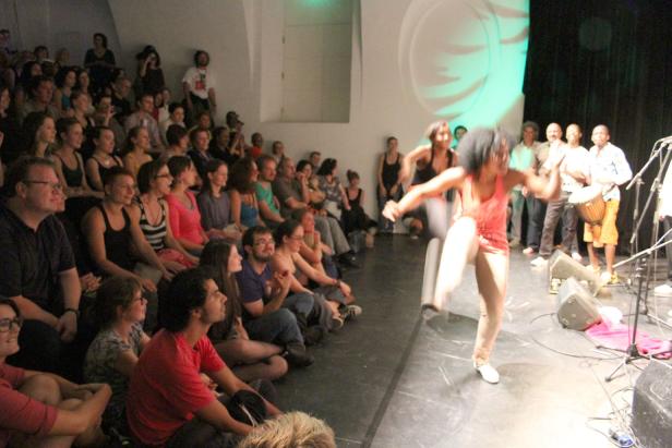 Eine Tänzerin führt vor einem sitzenden Publikum auf einer Bühne eine energiegeladene Performance auf.