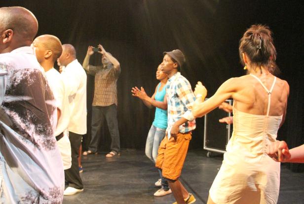 Eine Gruppe von Menschen tanzt auf einer Bühne vor dunklem Hintergrund.