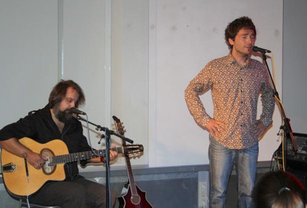 Ein Gitarrist spielt, während ein Sänger mit den Händen in den Hüften daneben steht.