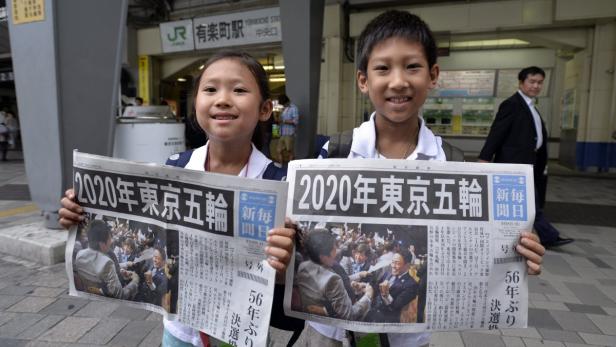 Zwei Kinder halten Zeitungen mit der Schlagzeile „Olympische Spiele 2020 in Tokio“ in die Kamera.