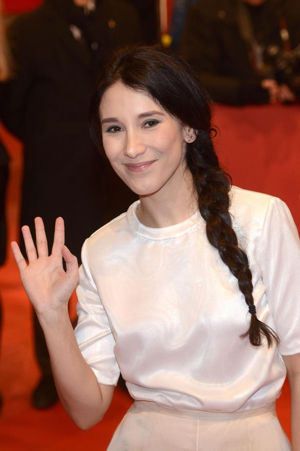 Eine Frau mit Zopf und hellem Oberteil winkt in die Kamera.