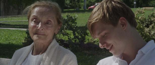 Eine ältere Frau und ein junger Mann sitzen zusammen auf einer Bank im Grünen.