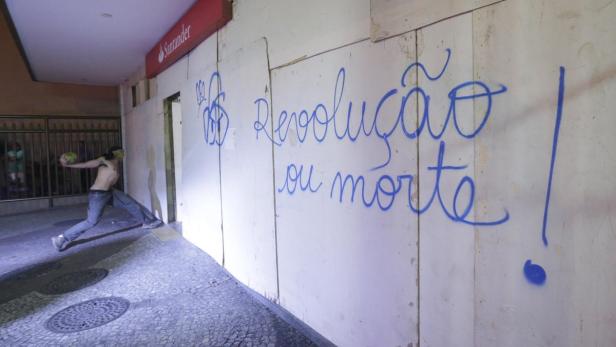 Ein Mann wirft einen Gegenstand vor eine Wand mit der Aufschrift „Revolução ou morte!“.