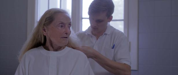 Eine ältere Frau im weißen Hemd wird von einem jungen Mann im weißen Poloshirt betreut.