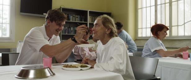 Ein junger Mann füttert eine ältere Dame in einem hellen Raum.