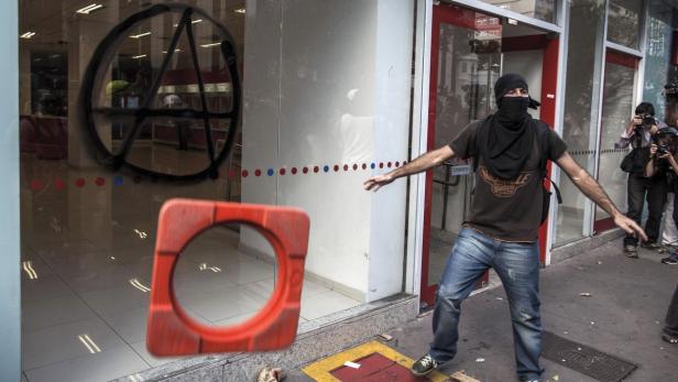 Ein Vermummter wirft einen Leitkegel gegen eine Schaufensterscheibe, die mit einem Anarchie-Symbol besprüht ist.