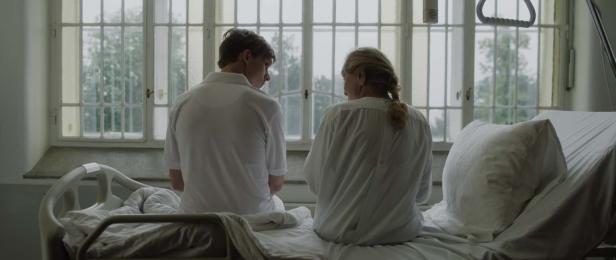 Ein junger Mann und eine ältere Frau sitzen auf einem Krankenbett und schauen aus dem Fenster.
