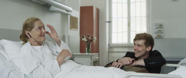 Eine ältere Frau liegt mit Kopfhörern im Krankenhausbett, während ein junger Mann sie anlächelt.