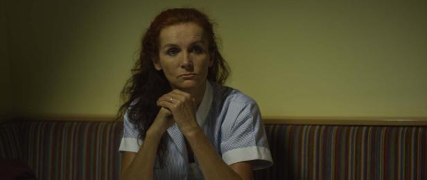 Eine Frau mit roten Haaren sitzt mit verschränkten Händen vor einer gelben Wand.