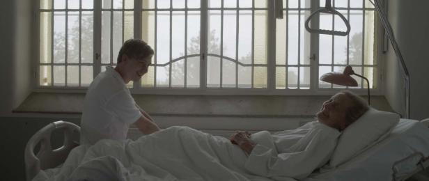 Ein junger Mann besucht eine ältere Frau, die in einem Krankenhausbett liegt.