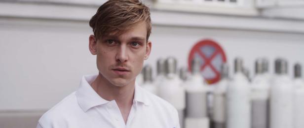 Ein junger Mann in weißem Poloshirt steht vor einem Gebäude mit Sauerstoffflaschen.