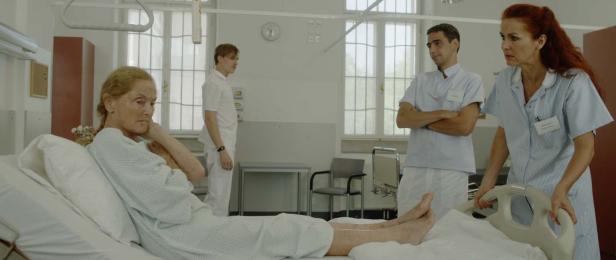 Eine ältere Frau liegt in einem Krankenhausbett, umgeben von medizinischem Personal.