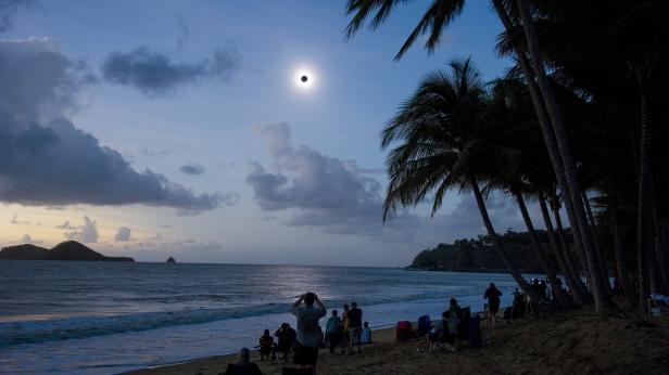 Menschen beobachten eine Sonnenfinsternis am Strand unter Palmen.