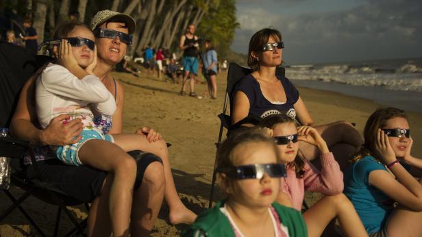 Mehrere Personen mit speziellen Brillen sitzen am Strand und schauen in den Himmel.