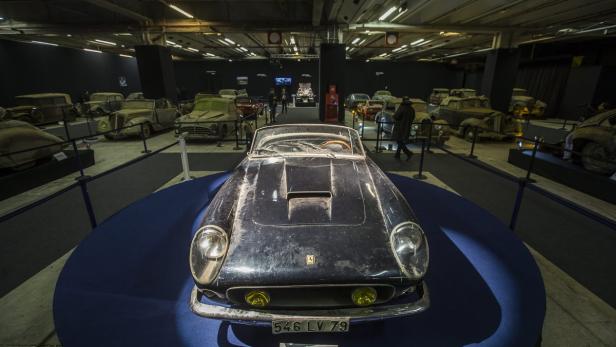 Ein abgenutzter, blauer Ferrari steht auf einer blauen Plattform in einer Ausstellungshalle.