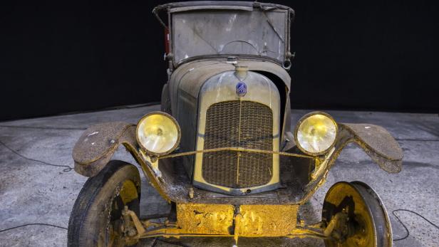 Ein verrosteter Oldtimer mit gelben Scheinwerfern steht vor einem dunklen Hintergrund.