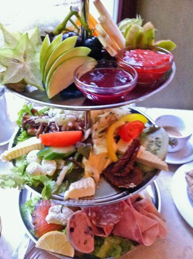 Ein dreistöckiger Servierständer gefüllt mit verschiedenen Speisen, darunter Obst, Käse und Wurstwaren.