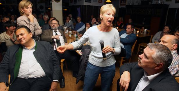 Eine Frau spricht mit einem Mikrofon vor einem Publikum in einem Raum.